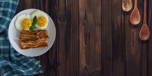Wood background with blue kitchen towel left and wooden kitchen spoons right. White plate with two fried eggs and Nature's Choice plant-based vegan bacon strips. 